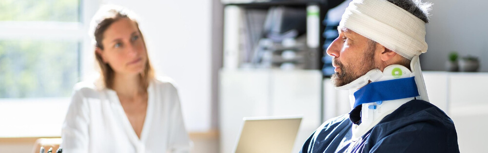 Unfallversicherung Ein Mann mit Kopfverband und Halskrause sitzt in einem Büro und spricht mit einer Frau, die ihm gegenüber sitzt.